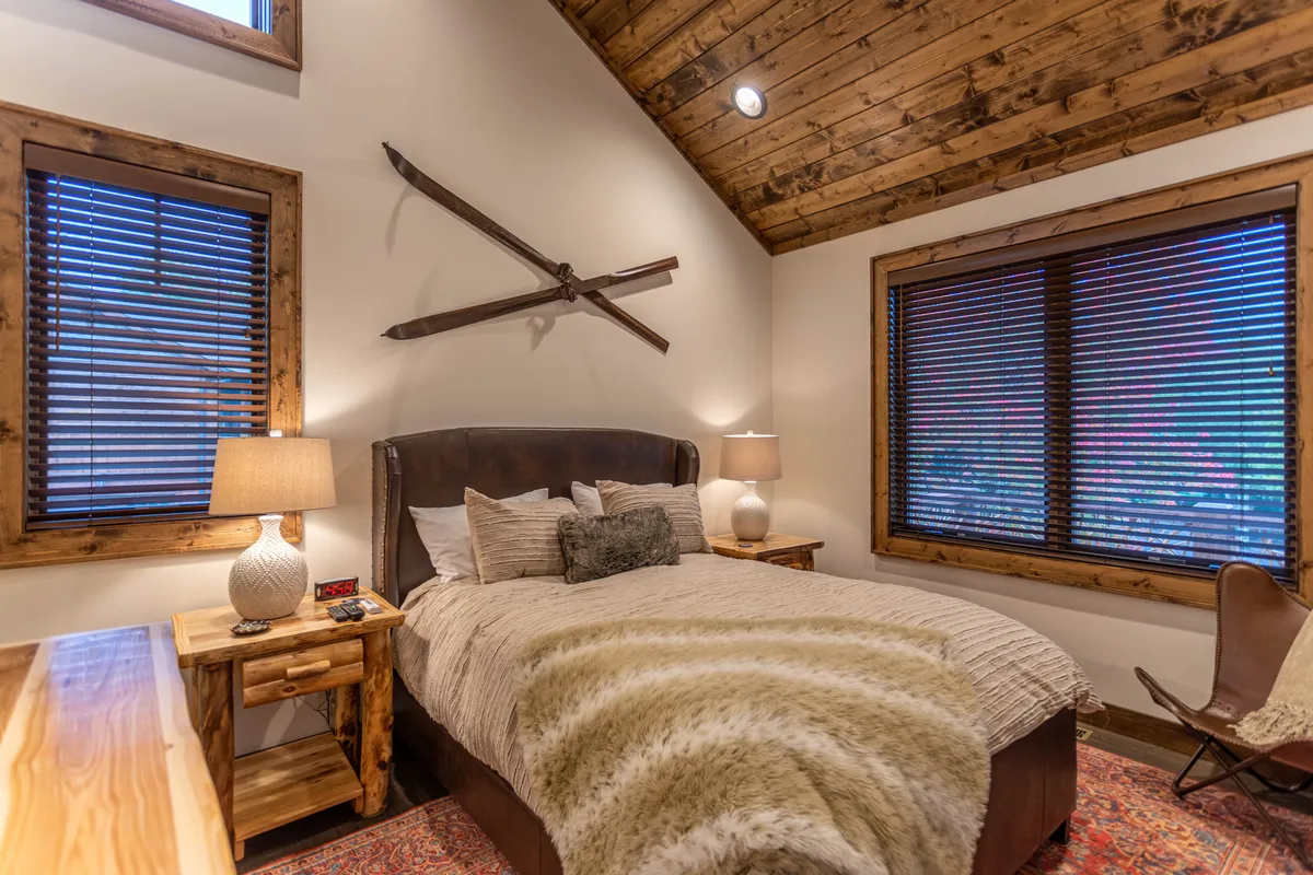 A tranquil bedroom featuring a comfortable bed with a plush duvet, nightstand with lamp, window letting in natural light, and decorative skis adding mountain charm. The palette is warm and restful, perfect for unwinding after a day of adventure.