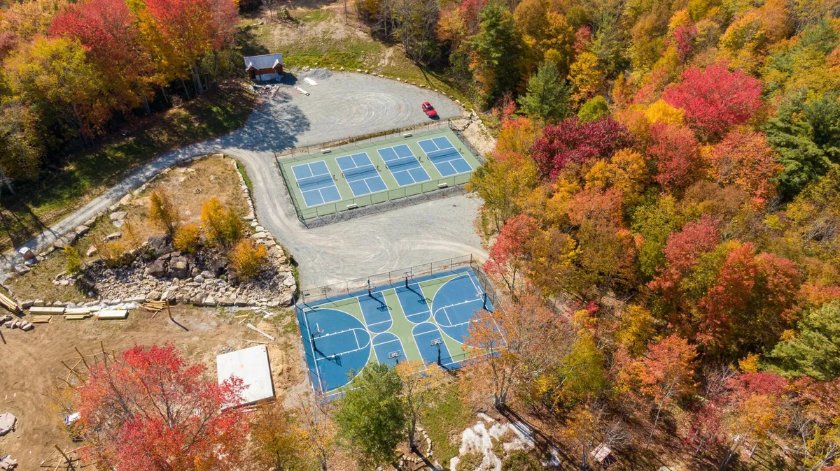 This vibrant exterior shot features tennis and basketball courts surrounded by colorful autumn foliage, with a parking area nearby. The courts are set against a backdrop of mature trees, suggesting a lively hub for sports and outdoor recreation within the community.