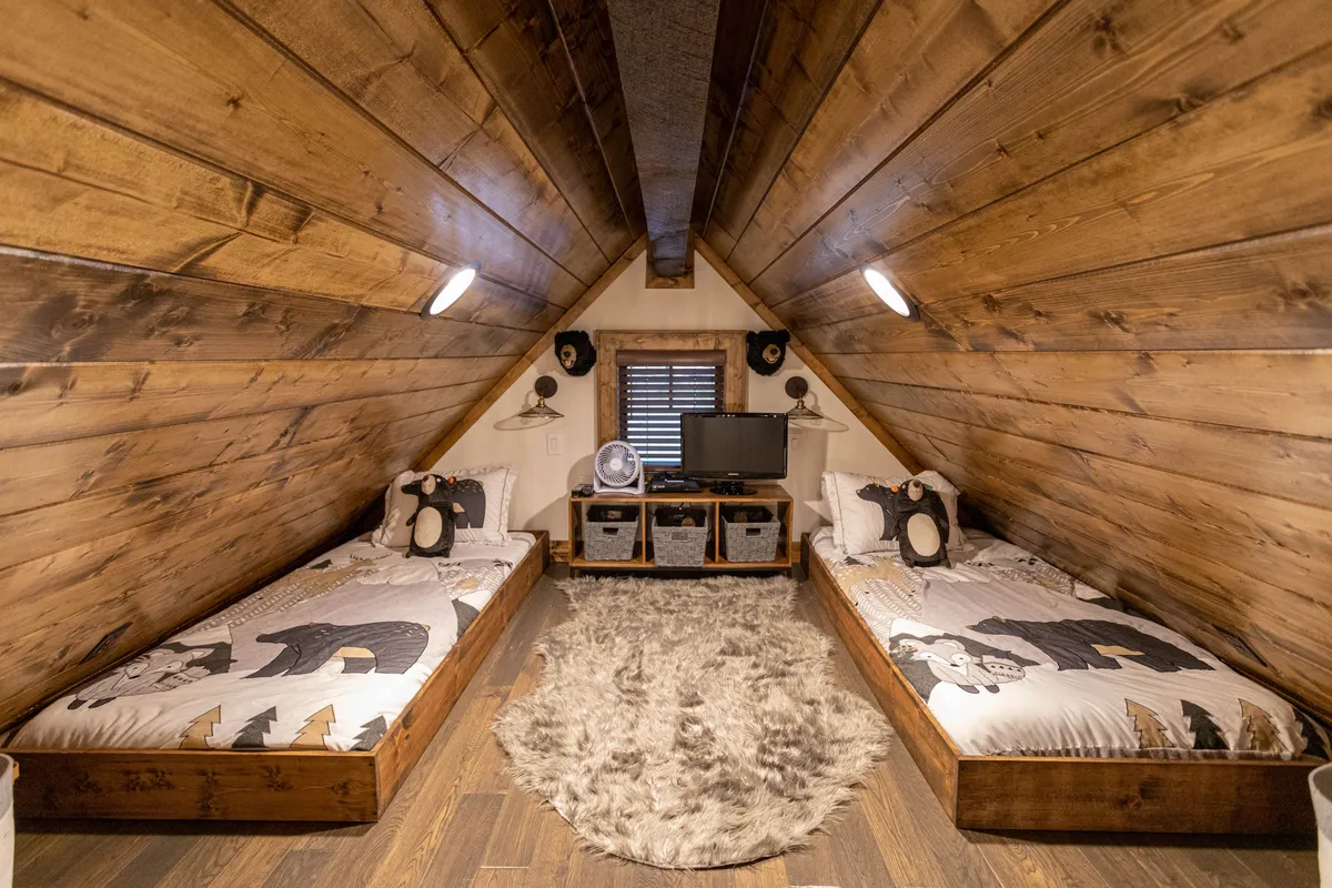 A warm, inviting loft space with twin beds featuring decorative pillows, a soft rug, wooden walls, and a television. The layout encourages both individual relaxation and shared moments, ideal for children or extra guests.