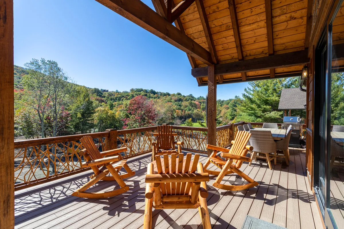 A spacious wooden deck furnished with rocking chairs and a dining set, overlooking a scenic view of trees. The area is bathed in natural light, inviting guests to relax, dine, or socialize while embracing the mountain air. The arrangement suggests a comfortable place for outdoor meals, morning coffee, or sunset gatherings.