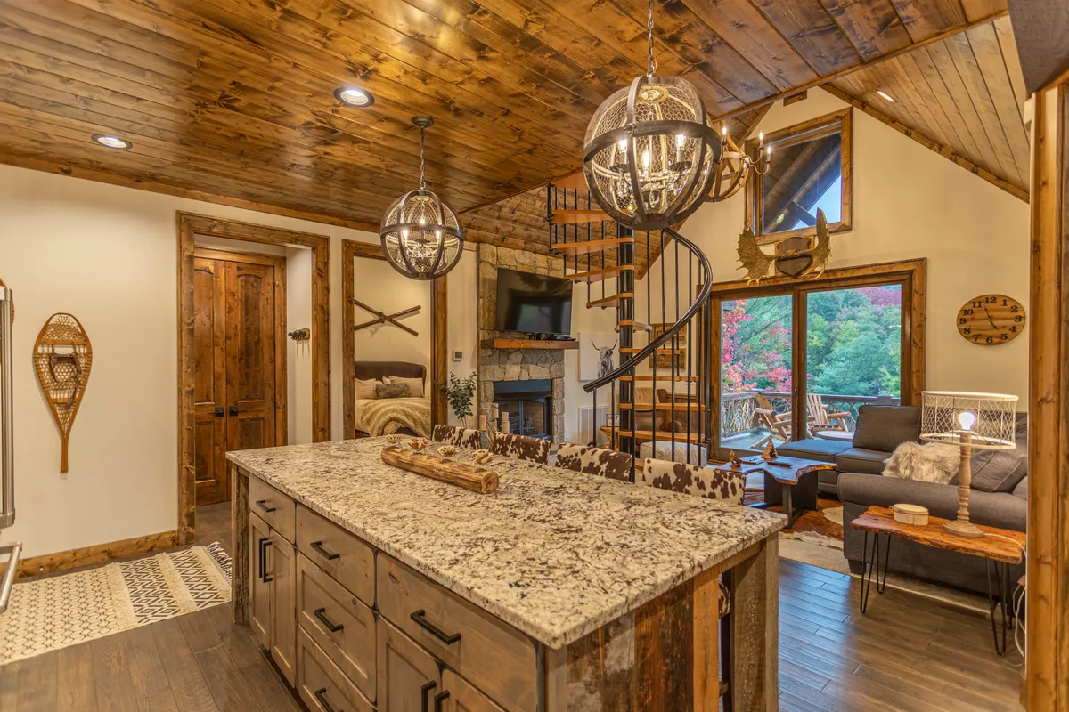 This image showcases the open flow between the granite kitchen island, the spiral staircase, and a cozy seating area. Large windows flood the open-concept space with natural light, providing a view of the outdoors. Plush sofas, the kitchen area, and transitions between living and dining spaces are all visible, reinforcing the sense of a connected gathering hub.