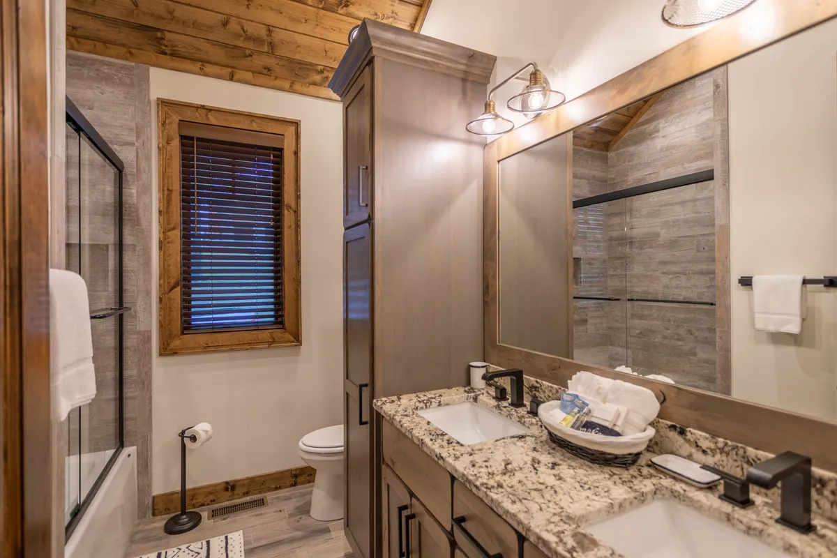 A spacious ensuite bathroom with double granite vanities, a large mirror, natural wood accents, and a glass-enclosed shower. Soft natural light fills the room, highlighting the spa-inspired fixtures and serene design.