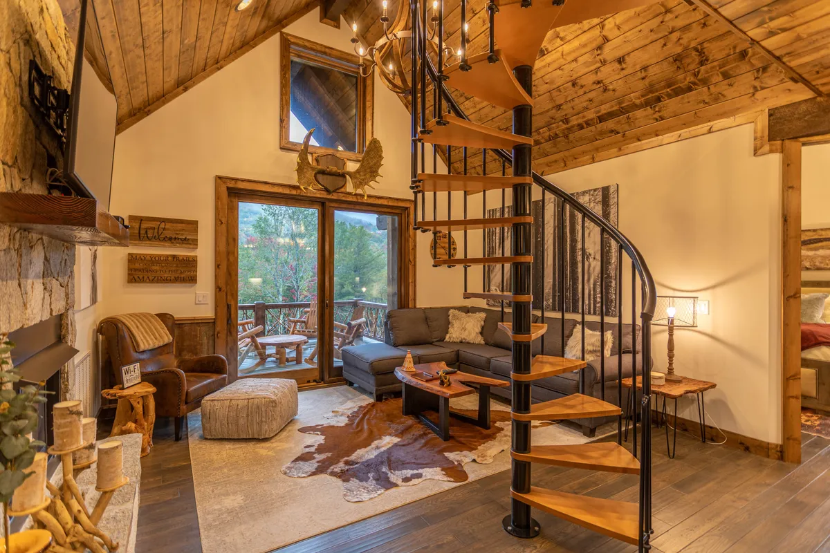 This inviting scene centers on the living area with a plush sofa, a glowing gas log fireplace, spiral staircase, and large windows. The arrangement suggests a space for relaxing, entertaining, and gathering around the fire after a day of adventure.