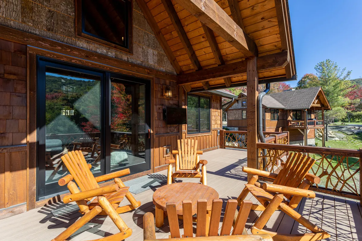 A cozy outdoor living area featuring wooden chairs and a round wooden table on a deck framed by large glass doors and overhead wooden beams. The space overlooks changing seasonal foliage, suggesting a perfect spot for wine tasting, sunset toasts, or relaxing after a day of adventure.