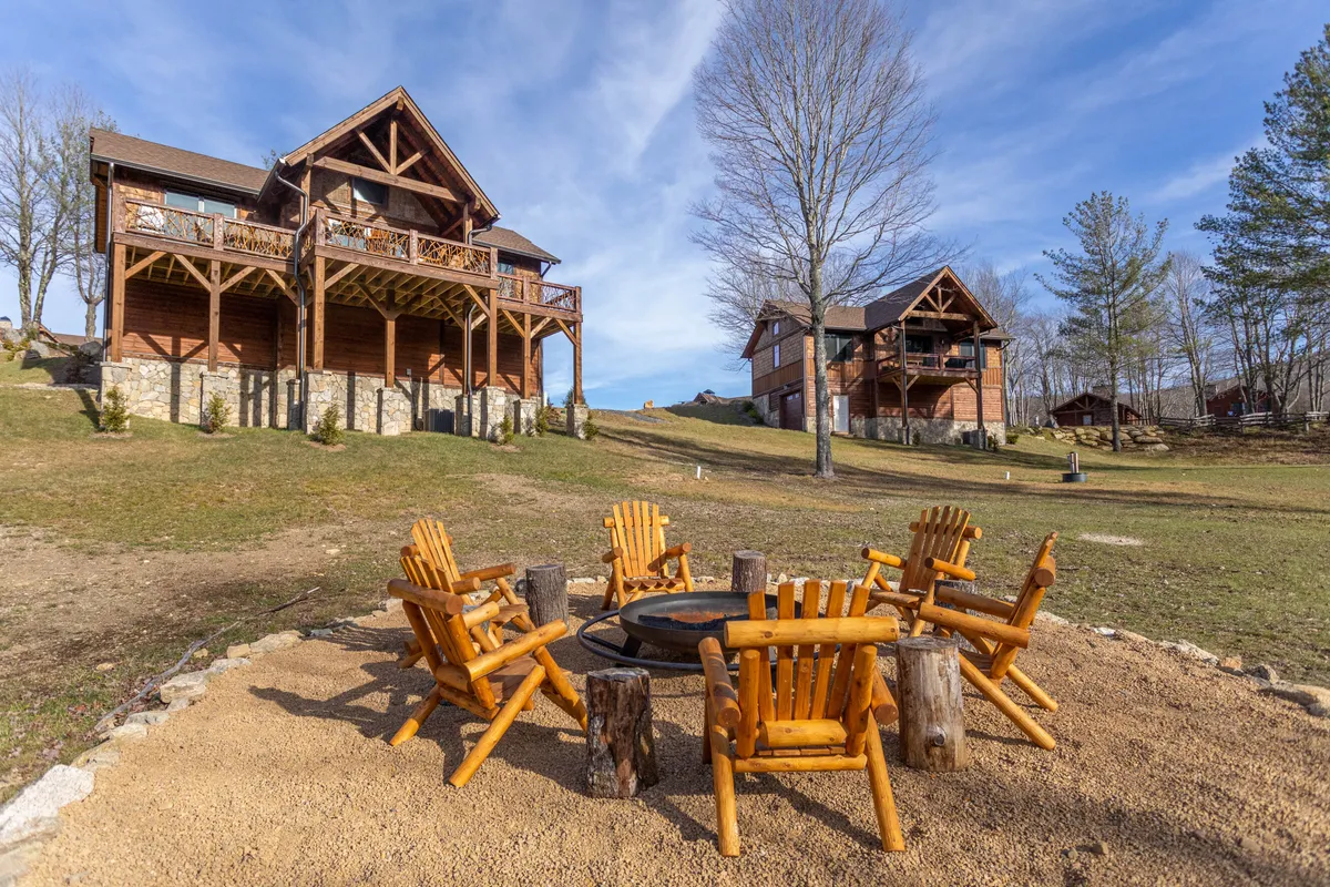 A cozy firepit area with wooden chairs arranged in a circle, set before a large wooden lodge and surrounded by greenery. The scene evokes a sense of warmth and togetherness, ideal for campfire gatherings, marshmallow roasting, and late-night conversations under the stars.