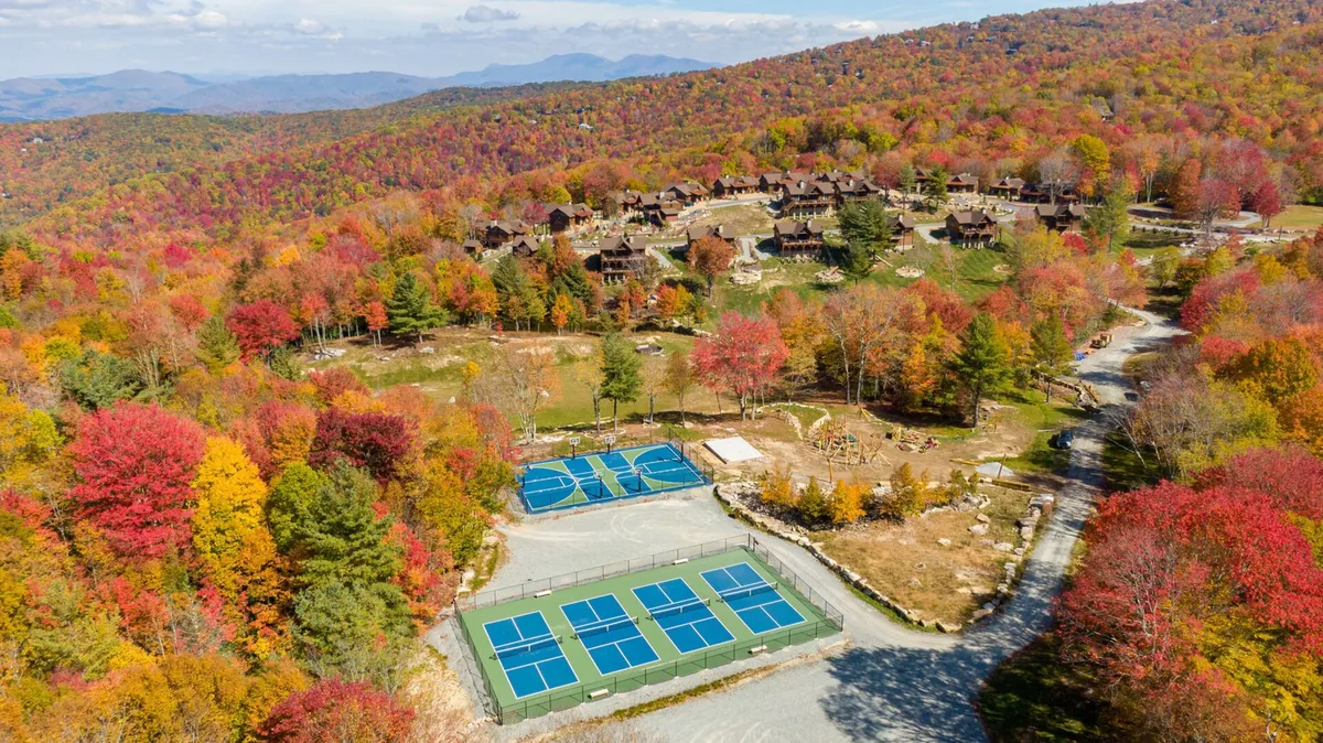 An exterior view of vibrant fall foliage surrounding inviting tennis courts and idyllic cabin exteriors. The scene is lively, suggesting active recreation (tennis, hiking, walks) and gatherings with family or friends against the backdrop of seasonal color.